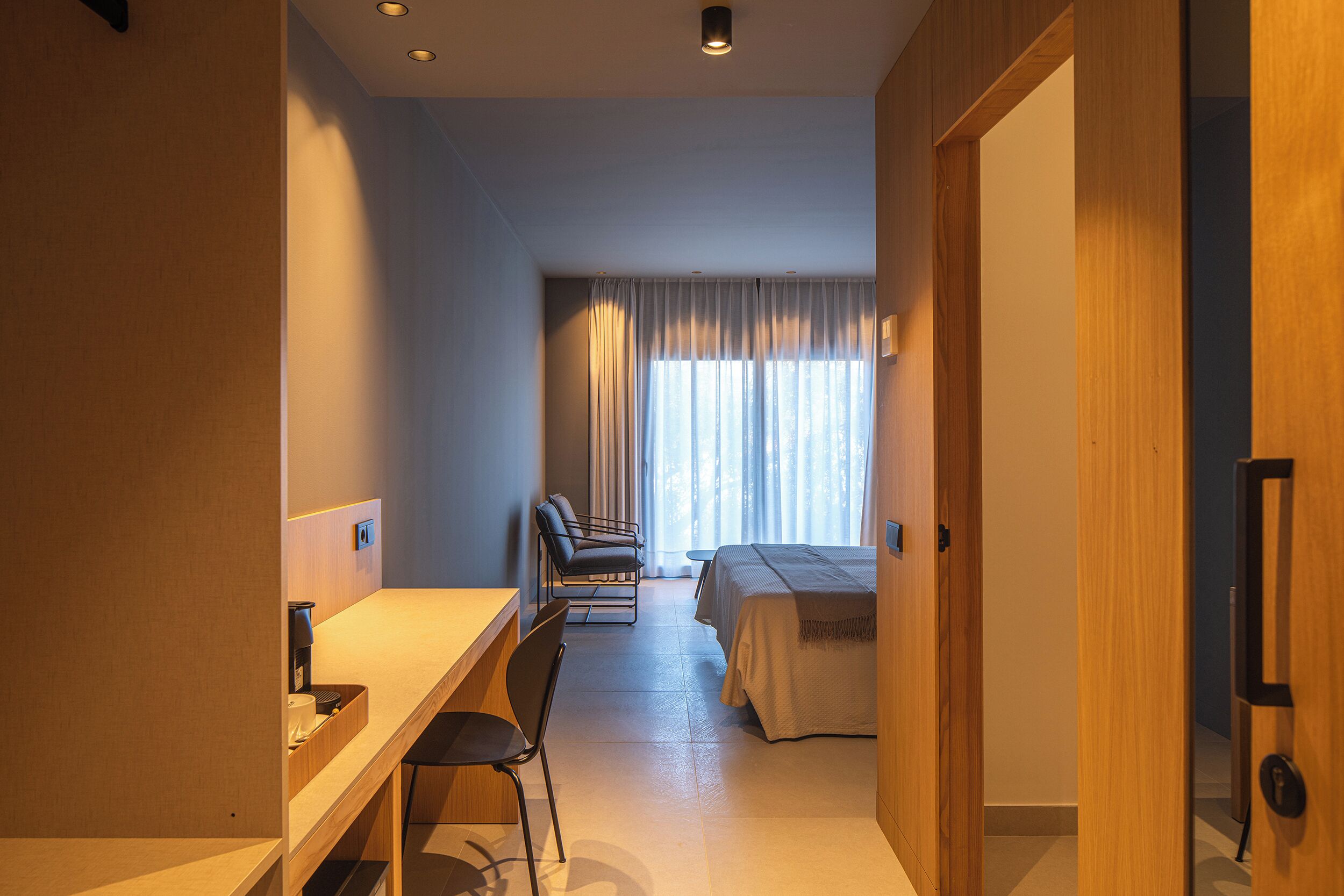 A hotel room with a desk, two chairs, a bed, and a window with curtains.