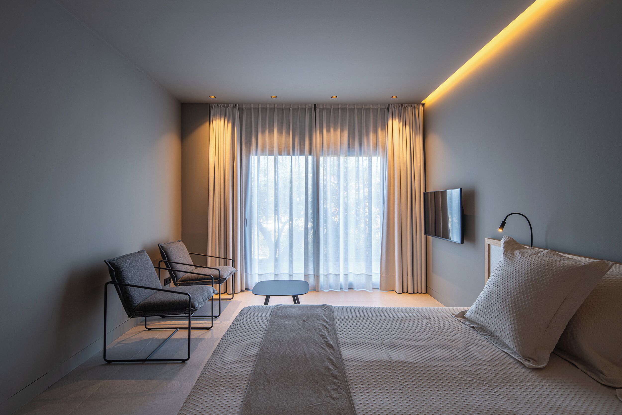 A modern hotel room with a bed, a seating area with chairs, a small table, and a TV mounted on the wall.