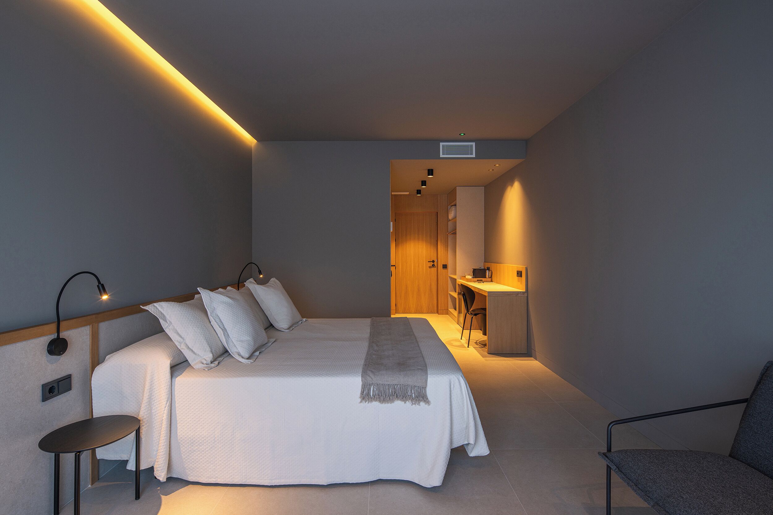 Un dormitorio con cama, mesita de noche, silla y un espacio de escritorio con otra silla.