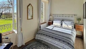 Een slaapkamer met een groot bed, een kroonluchter, een raam met gordijnen en uitzicht op een groen landschap.