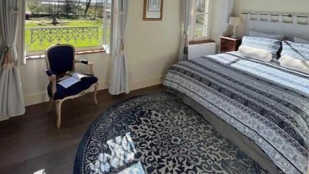 Een slaapkamer met een groot bed, een stoel, een rond tapijt en uitzicht op een tuin door een raam.