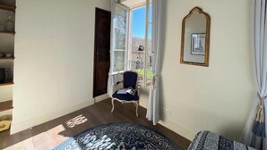 Een kamer met een groot raam, een stoel, een rond vloerkleed en een ingelijst schilderij aan de muur.