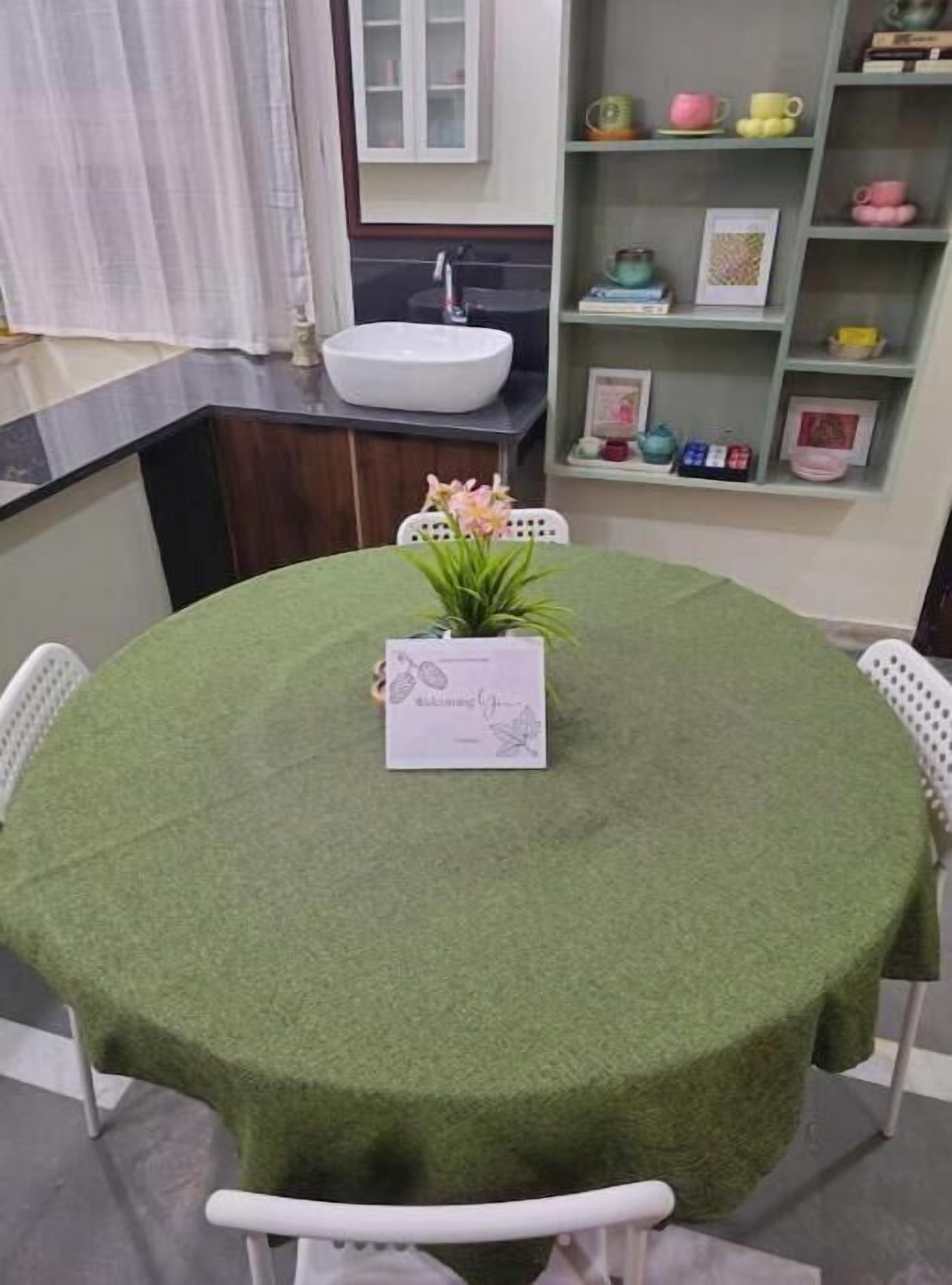 Un tavolo rotondo con tovaglia verde, una pianta in vaso e un cartoncino incorniciato.