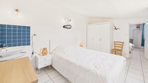 Un dormitorio con una cama, un armario blanco, una silla de madera, una mesita de noche con una lámpara y un baño con pared de azulejos azules y un lavabo blanco.
