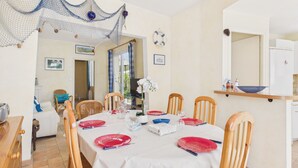 Comedor con mesa dispuesta para cuatro personas, sillas, televisor y una zona de cocina con refrigerador.