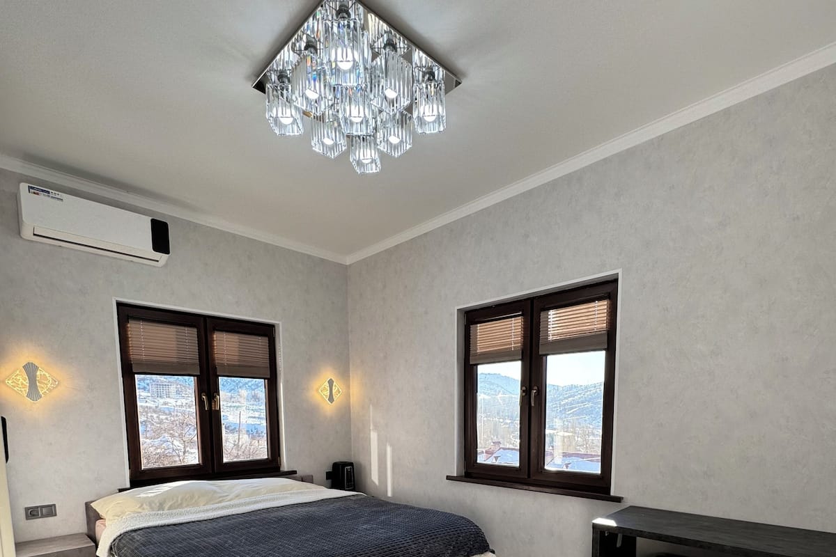 A bedroom with a bed, a desk, a chair, and a view of the city through the windows.