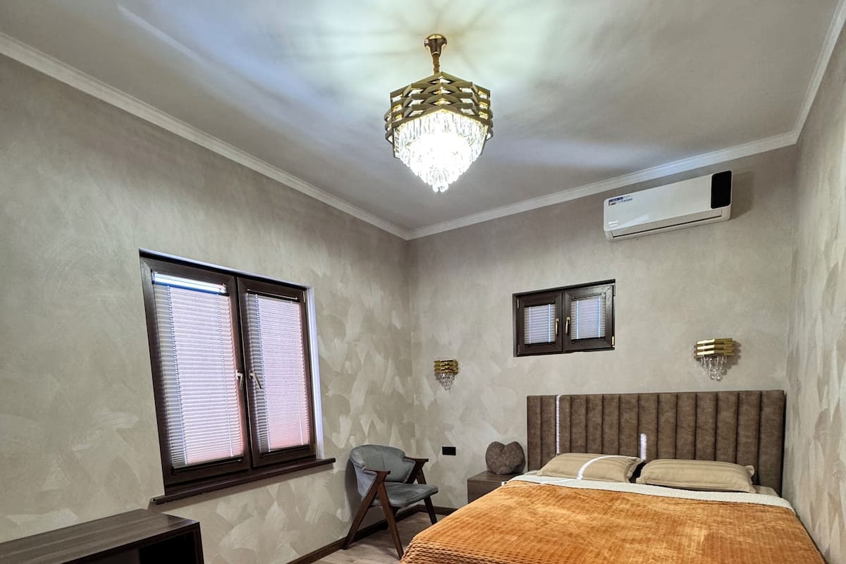 A bedroom with a bed, bedside tables, a chair, a window with blinds, an air conditioning unit, and a chandelier.