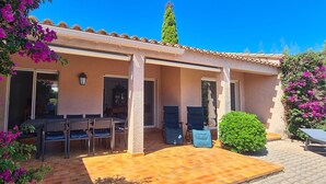 Eine überdachte Terrasse mit Tisch und Stühlen, ein kleiner Garten und ein Gebäude mit einem Ziegeldach.