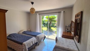 Ein Schlafzimmer mit zwei Betten, einem Holzschrank, einem kleinen Tisch und Blick auf einen Garten durch eine Schiebetür.
