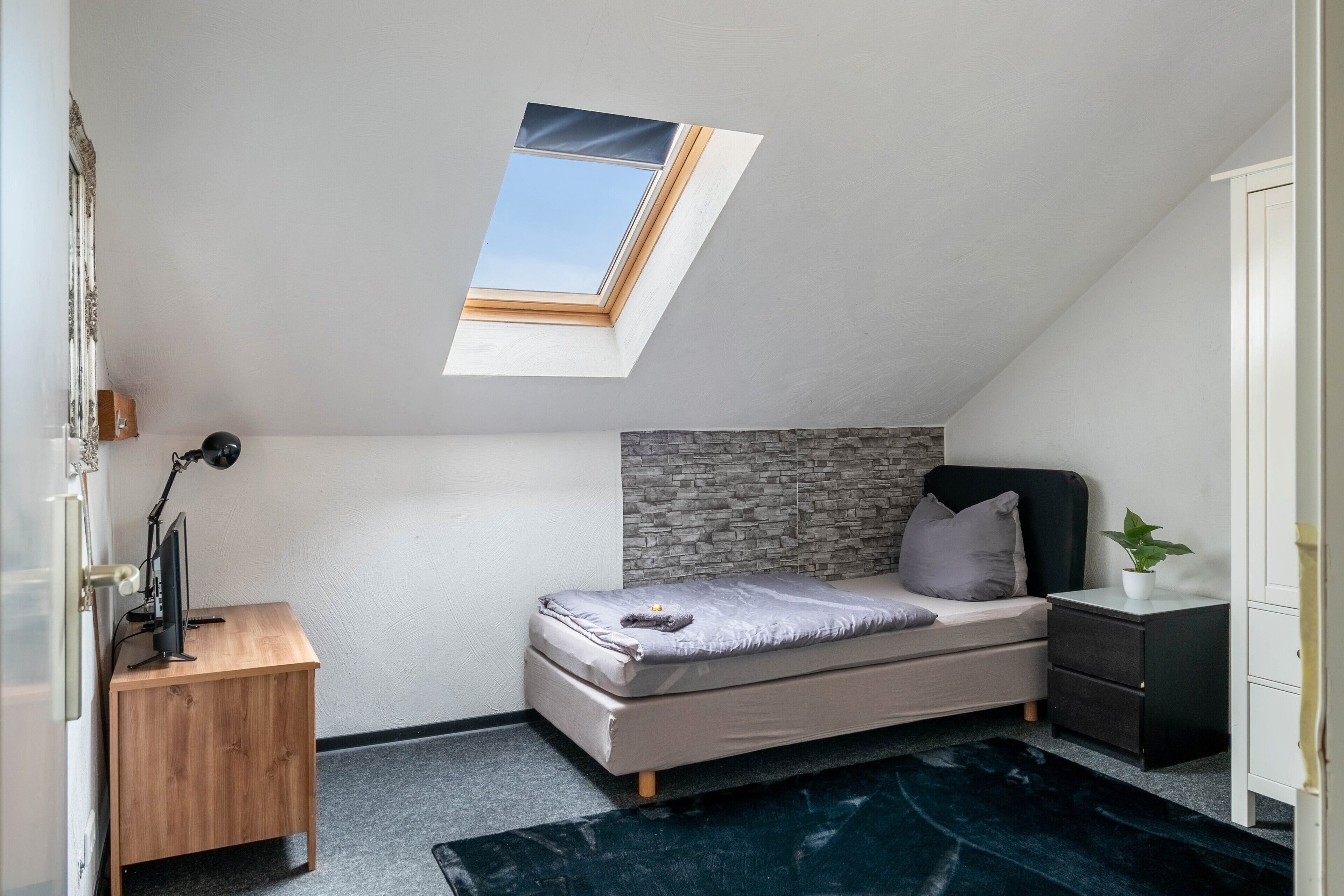 A bedroom with a sloped ceiling, a skylight, a bed, a desk, a computer, a plant, and a wardrobe.