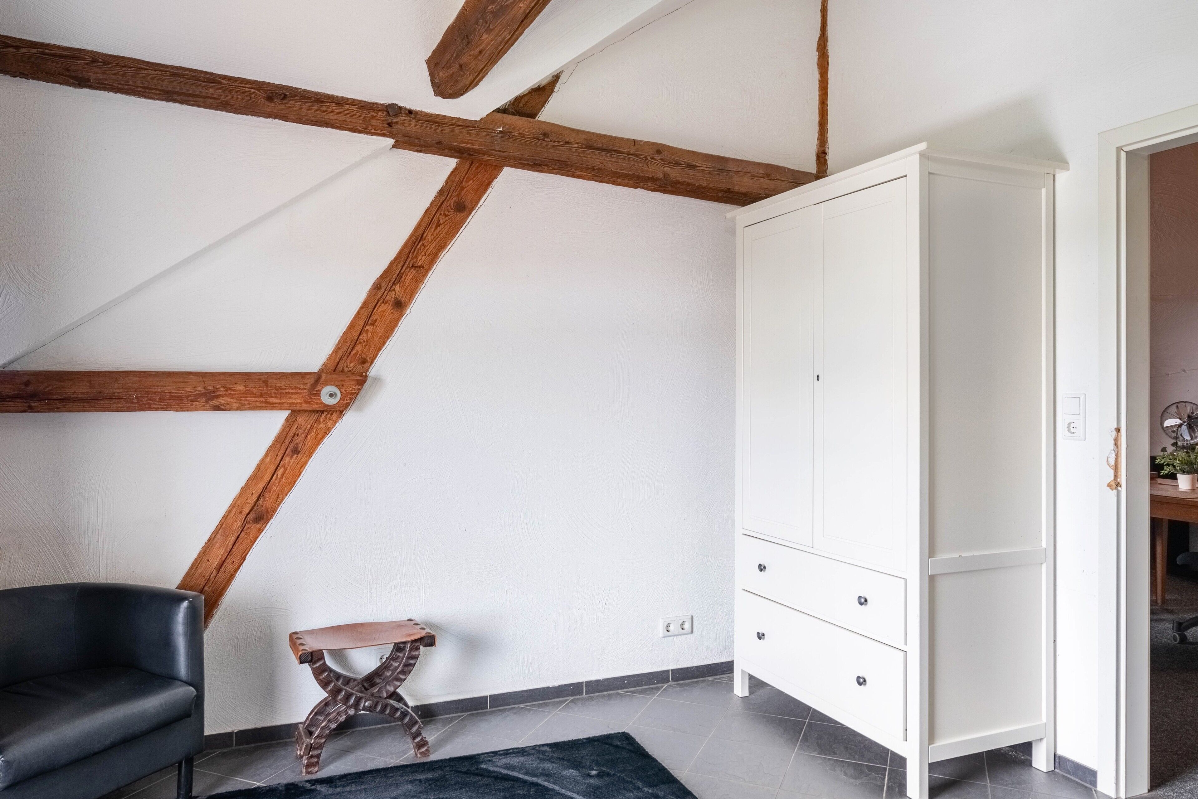 Une pièce avec des poutres apparentes, une armoire blanche, un fauteuil en cuir noir et un petit tabouret.