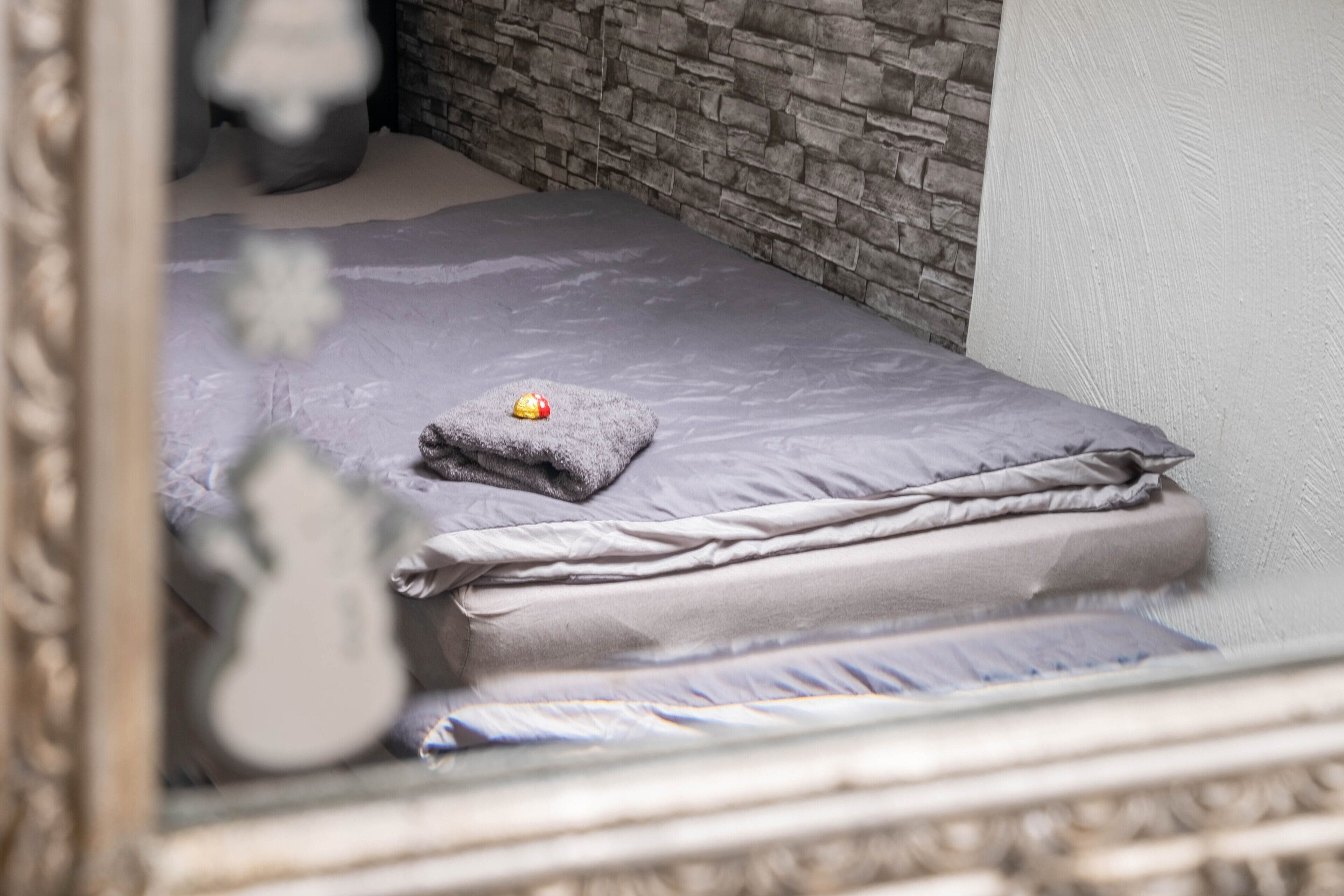 A small, neatly made bed with a grey mattress and pillows, a stone wall behind it, and a white wall to the right.