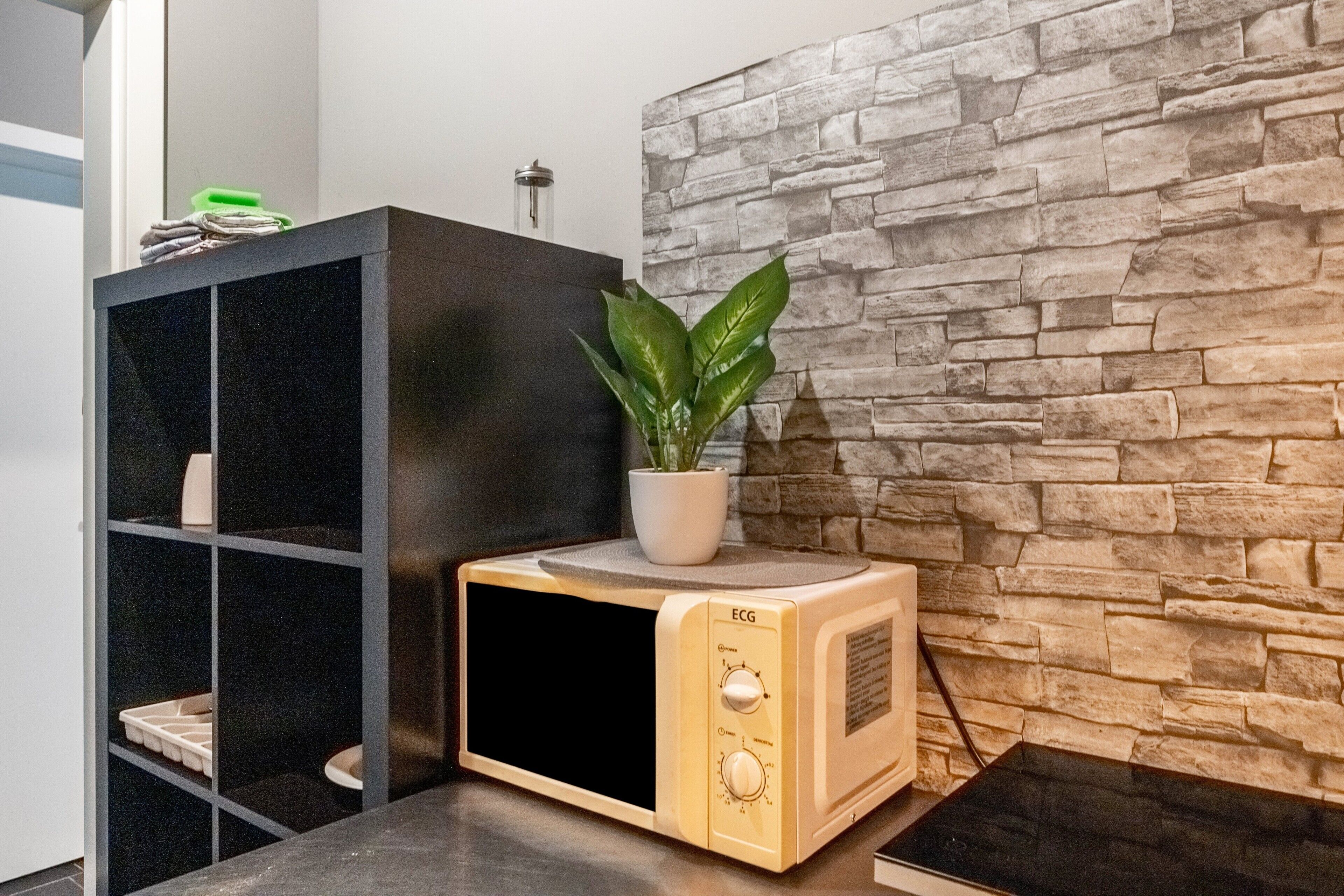 A microwave oven with a plant on top, a black shelving unit, and a stone-textured wall.