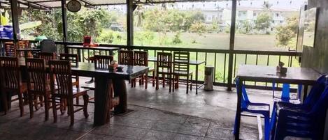 A covered outdoor seating area with tables and chairs, a tiled floor, and a view of greenery and buildings in the distance.