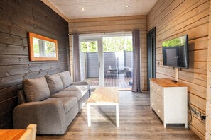 A cozy cabin interior with a sofa, a wooden coffee table, a TV mounted on the wall, and a sliding glass door leading outside. - Pine Lodge Spa Pet (Thirsk)
