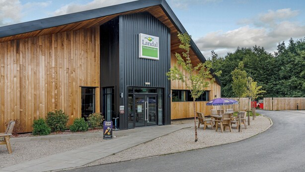 A building with a sign reading 'Landal Woodland Lake,' a wooden exterior, and outdoor seating with a table and chairs. - Pine Lodge Spa Pet (Thirsk)