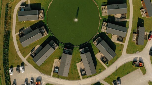 An aerial view of a circular building complex with a central pond, surrounded by multiple buildings with flat roofs, arranged in a radial pattern. - Pine Lodge Spa Pet (Thirsk)