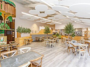 A restaurant interior with round tables, cushioned seating, and a kitchen area in the background. - Pine Lodge Spa Pet (Thirsk)