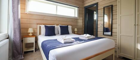 A bedroom with a wooden paneled wall, a large bed with blue bedding, a nightstand with a lamp, a wardrobe, and a window with curtains.