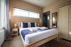 A bedroom with a wooden paneled wall, a large bed with blue bedding, a nightstand with a lamp, a wardrobe, and a window with curtains. - Pine Lodge Spa Pet (Thirsk)