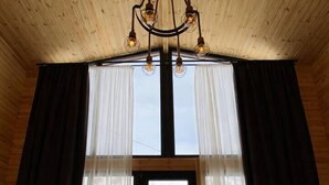 Ein Zimmer mit Holzwänden und -decke, einem großen Fenster mit transparenten Vorhängen sowie einem Kronleuchter.