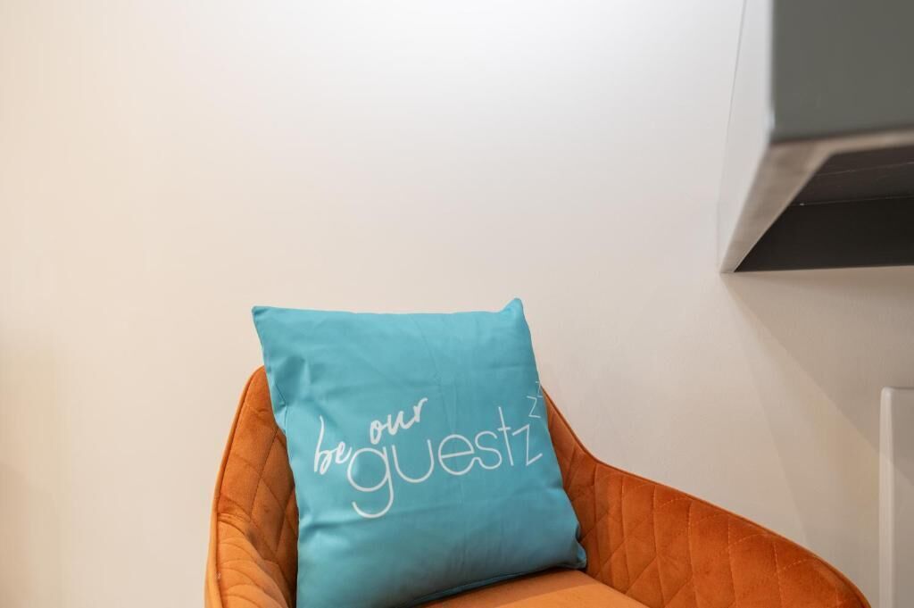 A chair with a teal pillow that reads 'be our guestz'.