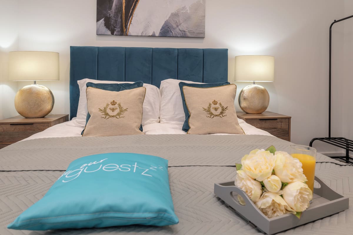 Habitación de hotel con una cama, dos lámparas de noche, un cojín decorativo, una bandeja con flores y un vaso de zumo de naranja.