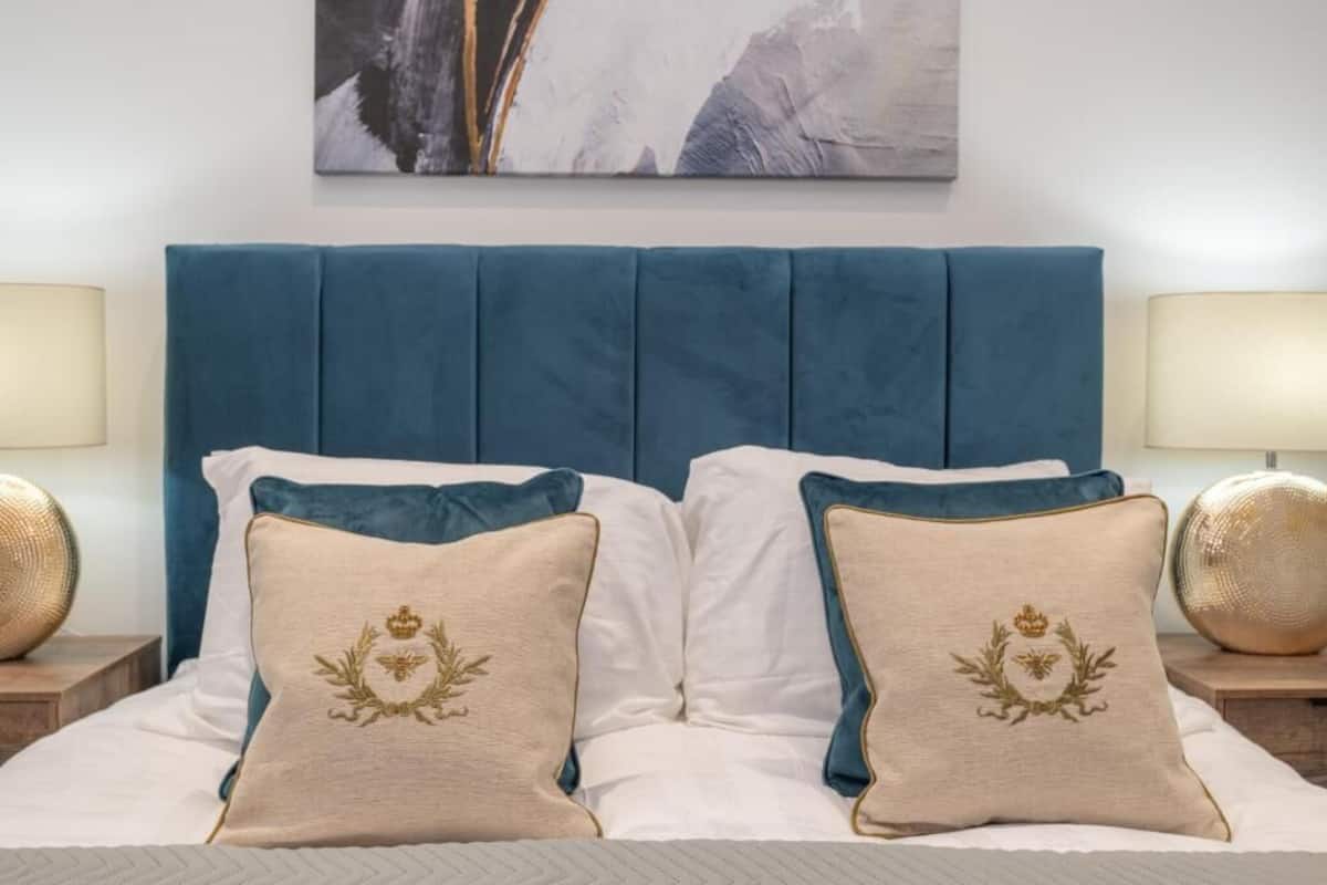 Una cama con cabecera azul, dos almohadones decorativos con diseño de cresta y un cuadro abstracto enmarcado encima.