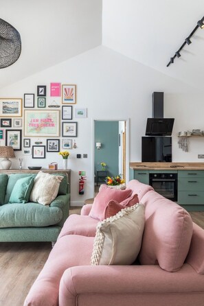 Un salon moderne avec un canapé vert, un canapé rose et un coin cuisine doté d’une hotte noire.