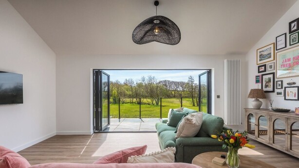 A modern living room with a green sofa, pink cushions, a wooden coffee table, and a view through a glass door to a green landscape.