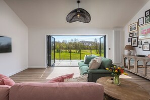 A modern living room with a green sofa, pink cushions, a wooden coffee table, and a view through a glass door to a green landscape.