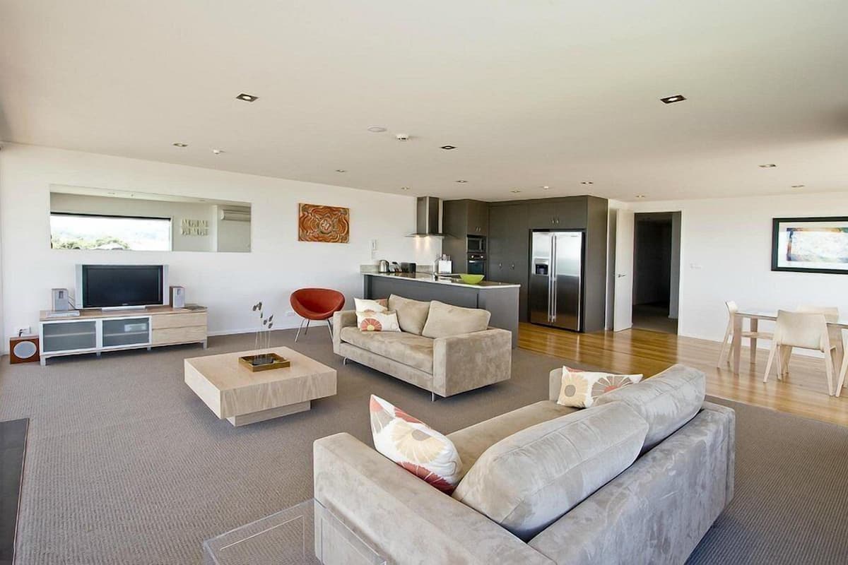 A modern living room with a flat-screen TV, a red chair, a grey sofa, and a kitchen area with a fridge and stove.