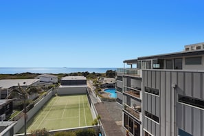 Un edifici modern amb una pista de tennis i una piscina, amb vista a un cel blau clar i al mar.