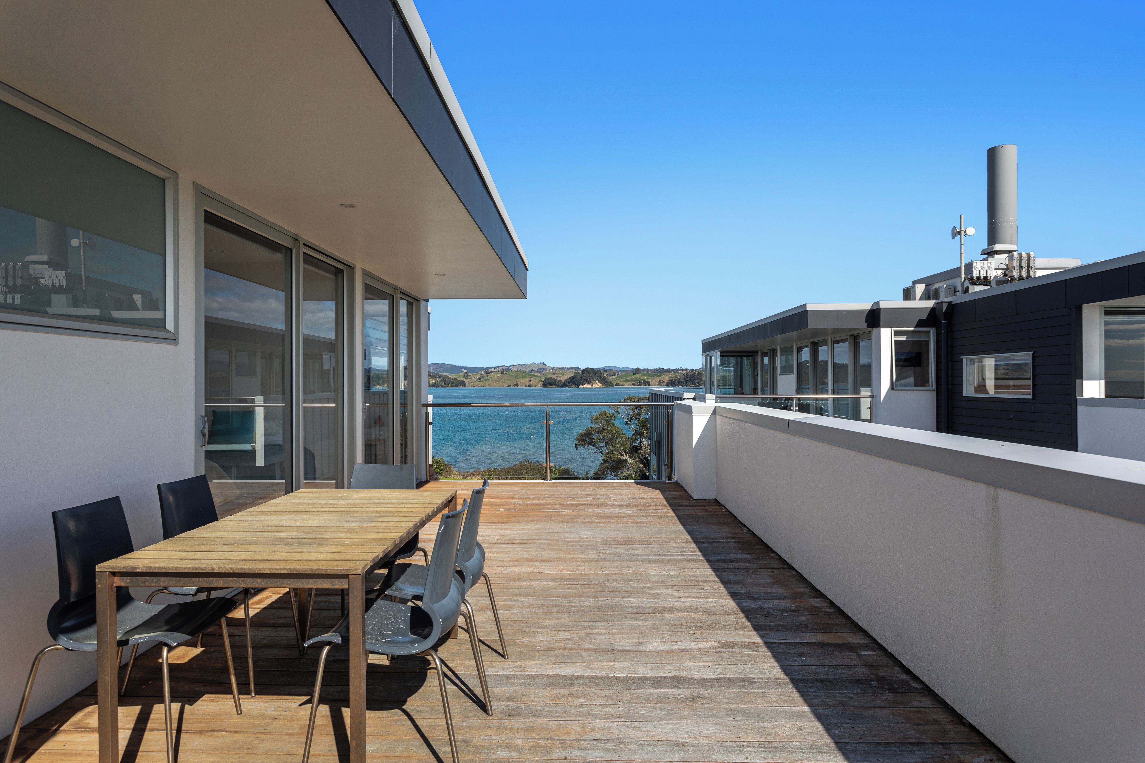 A modern outdoor dining area with a wooden table and chairs, overlooking a body of water and hills.
