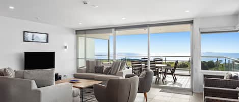 A modern living room with a flat-screen TV, grey sofas, a coffee table, and a view of the sea through large windows.