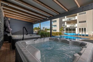 A covered outdoor area with two hot tubs, a pool, and a view of a modern building with balconies.