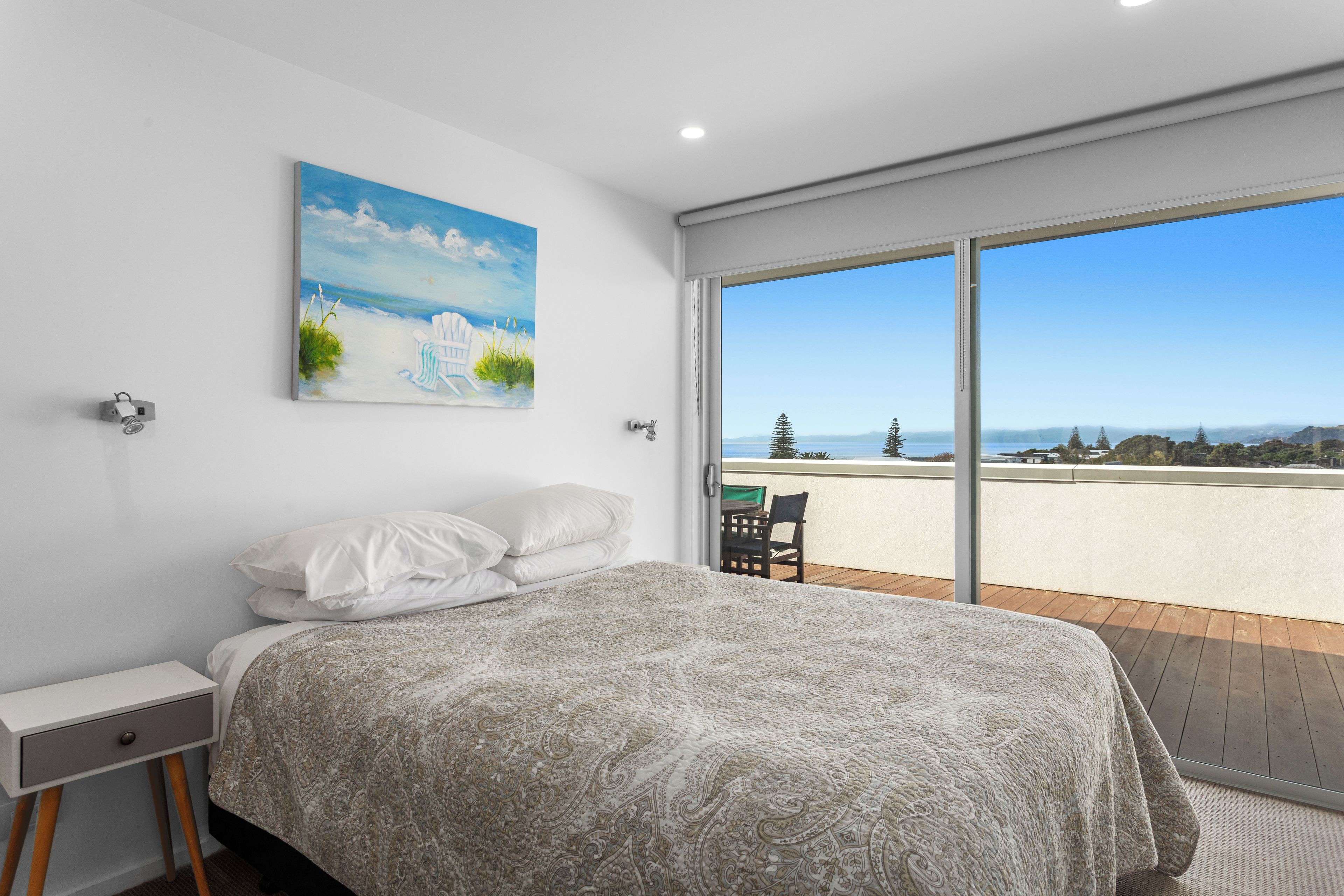 A bedroom with a bed, a nightstand, a painting, and a view of the outdoors through a large window.