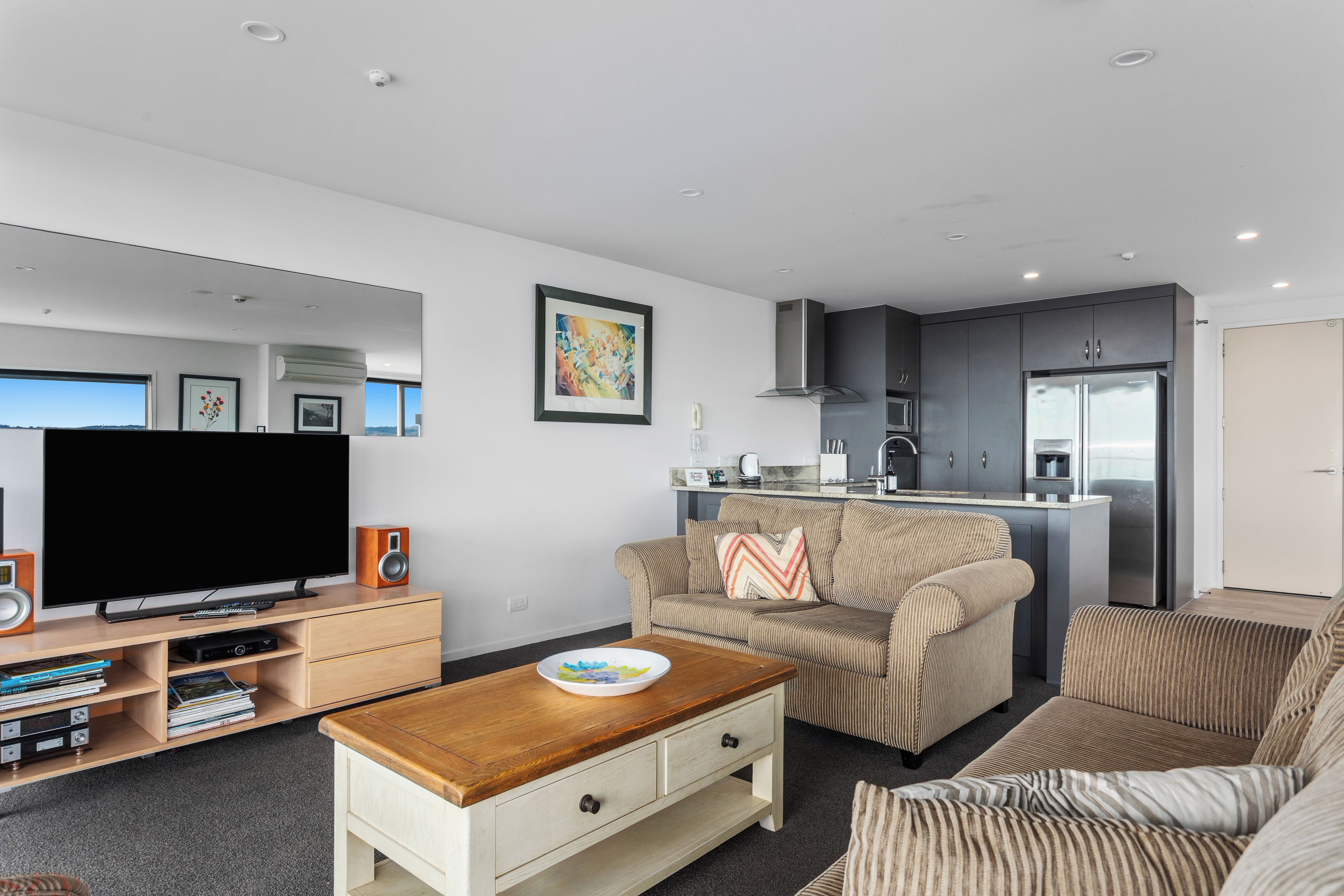 A modern living room with a flat-screen TV, a coffee table, and a sofa. There is a kitchen area with a refrigerator and a microwave.