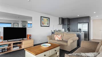 A modern living room with a flat-screen TV, a coffee table, and a sofa. There is a kitchen area with a refrigerator and a microwave.