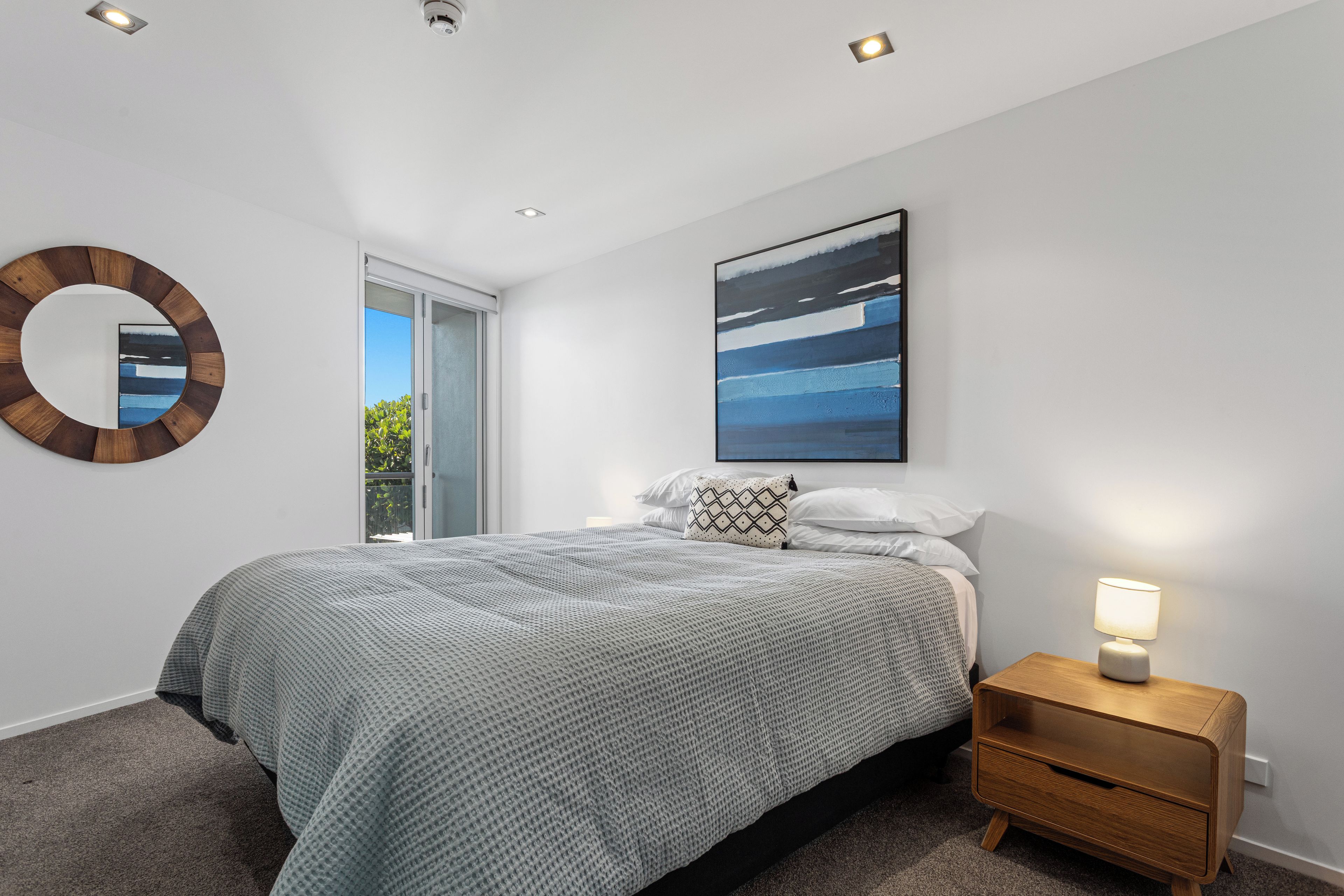 A bedroom with a bed, a bedside table, a nightstand, a wall-mounted mirror, and a painting.