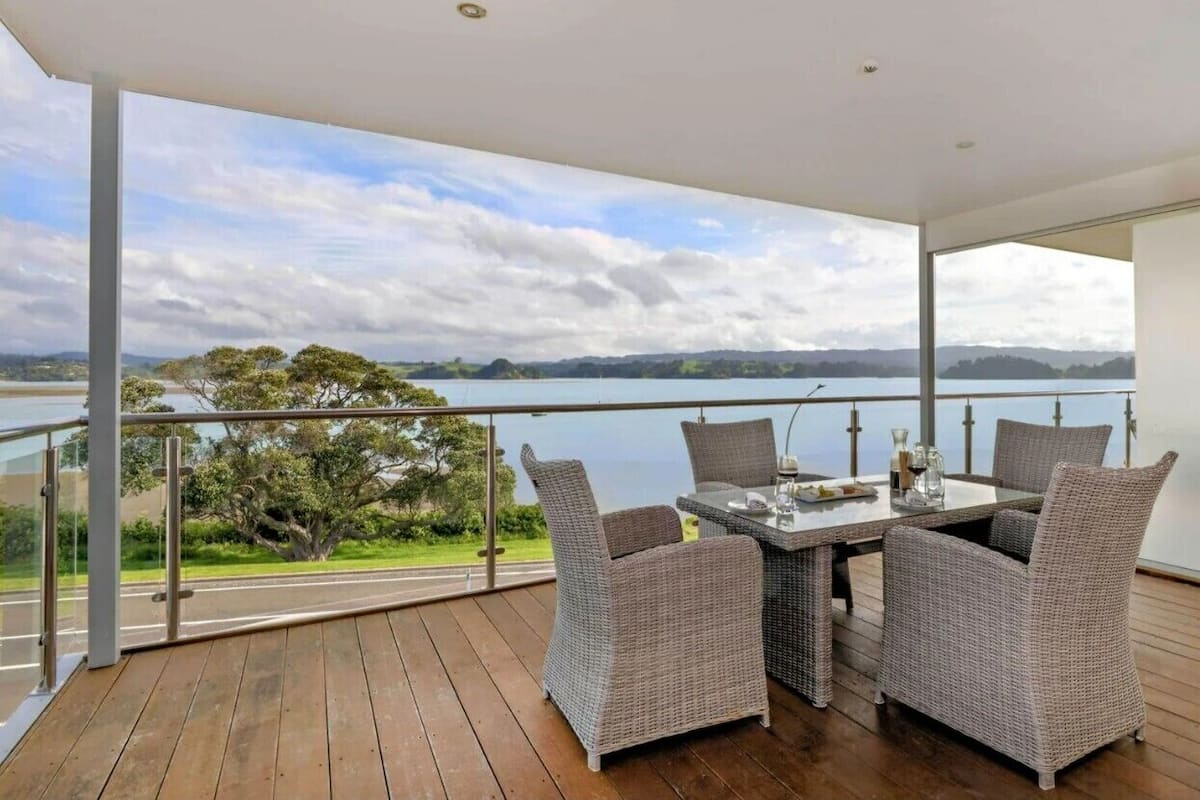 A balcony with wicker furniture, a glass table, and a view of a lake and distant hills.