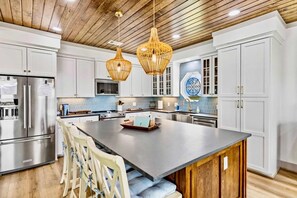 A modern kitchen with white cabinetry, a large island, hanging wicker pendant lights, and a stainless steel refrigerator. - Spoonbill at West Bay (Galveston)