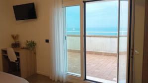 A room with a bed, a desk, a TV, and a sliding glass door to a balcony with a view of the sea.
