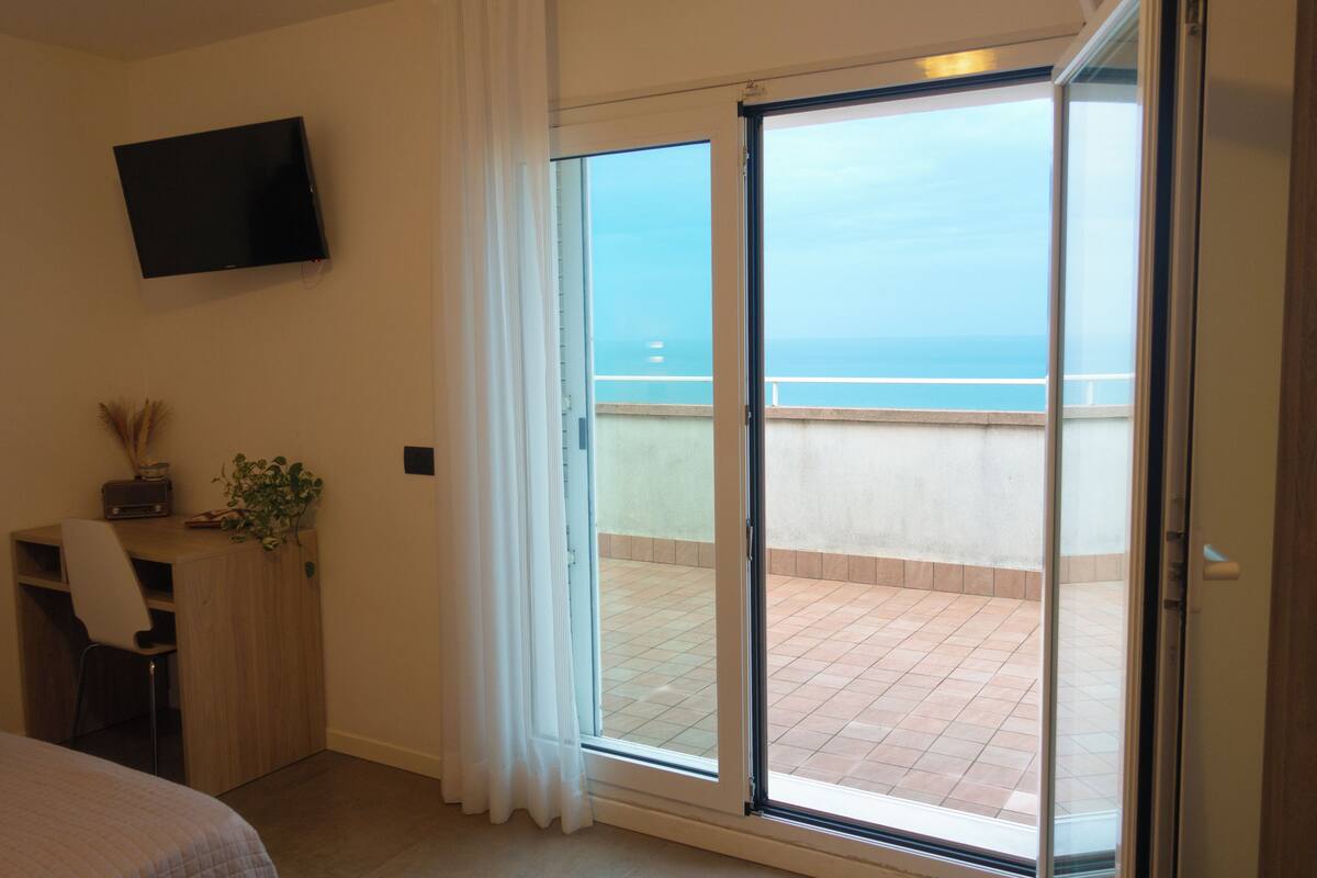 Una habitación con cama, escritorio, televisor y puerta corrediza de vidrio a un balcón con vista al mar.