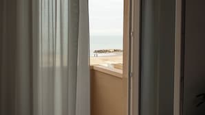 A view through a window with curtains, revealing a balcony with a view of a beach and rocky outcrops.