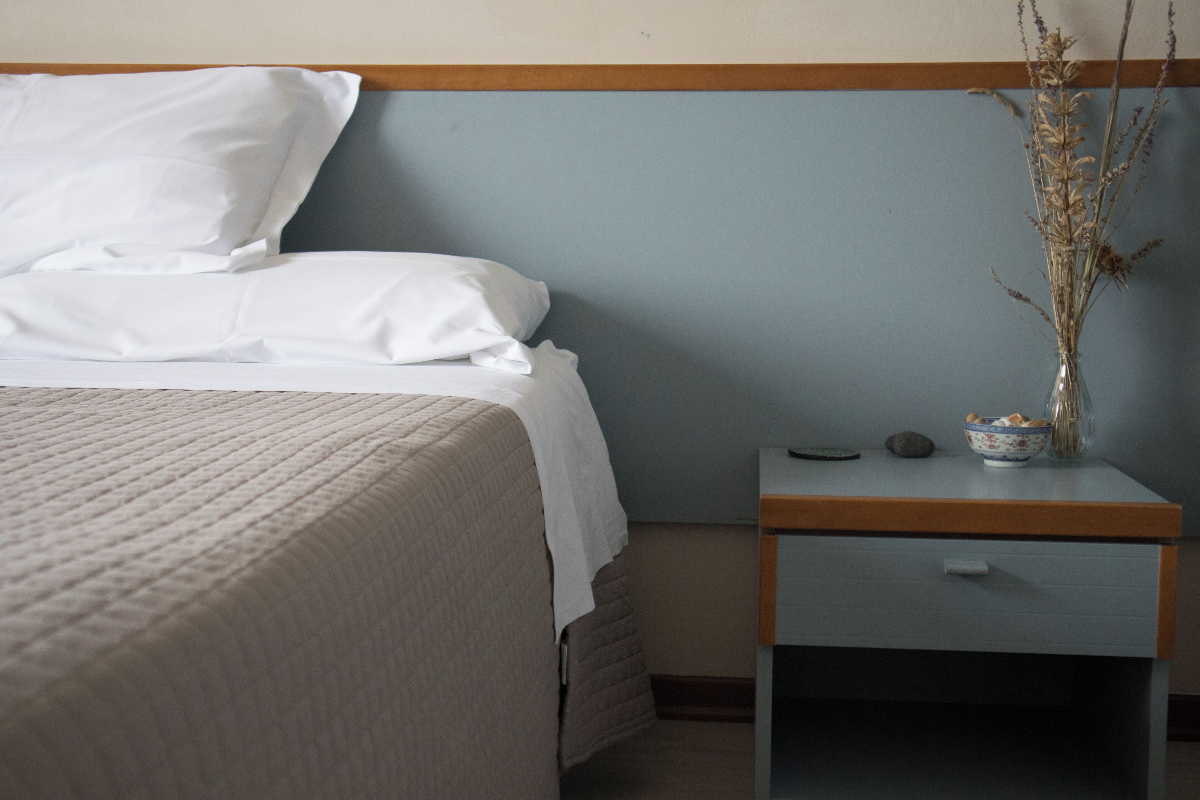 Una cama bien hecha con sábanas blancas, una mesita de noche con un cuenco y un jarrón de flores secas, y un cabecero de madera.