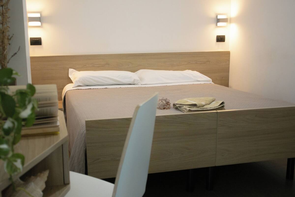 Una cama bien tendida con cabecera de madera, lámparas de noche y una mesita con una planta y algunos objetos encima.