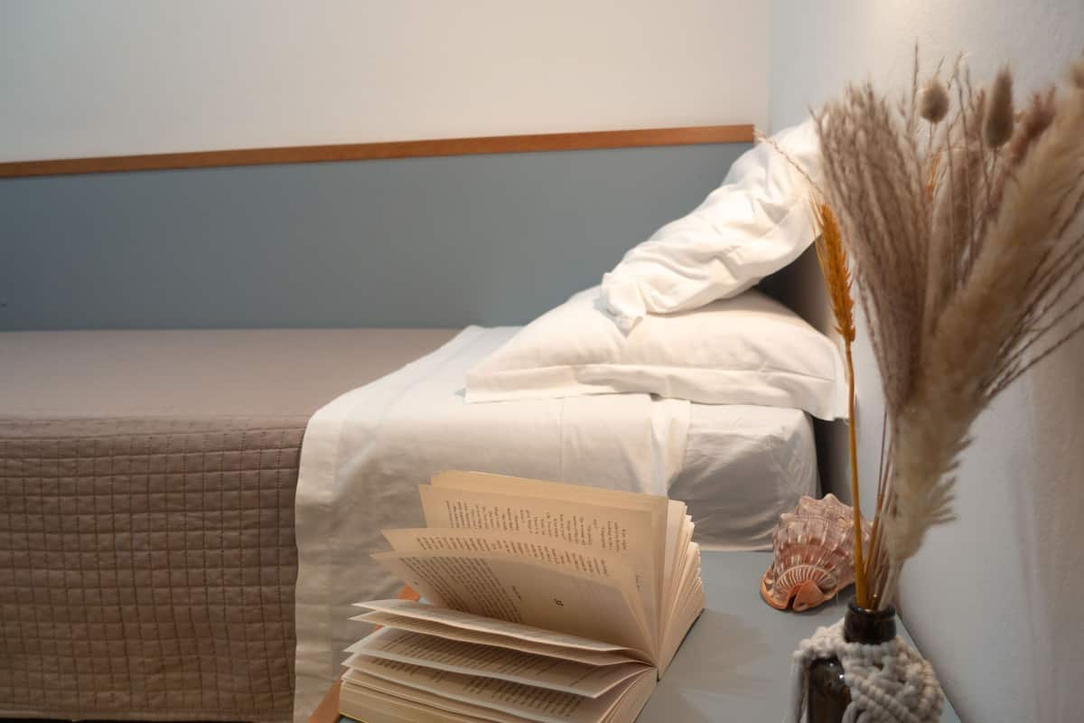 Una cama bien hecha con un edredón acolchado, una pila de libros y un florero decorativo con plantas secas y una concha marina.