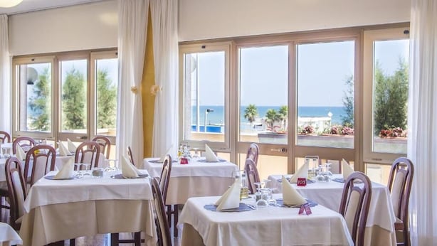 A dining area with tables set for guests, large windows with a view of the sea, and a curtain partially drawn.