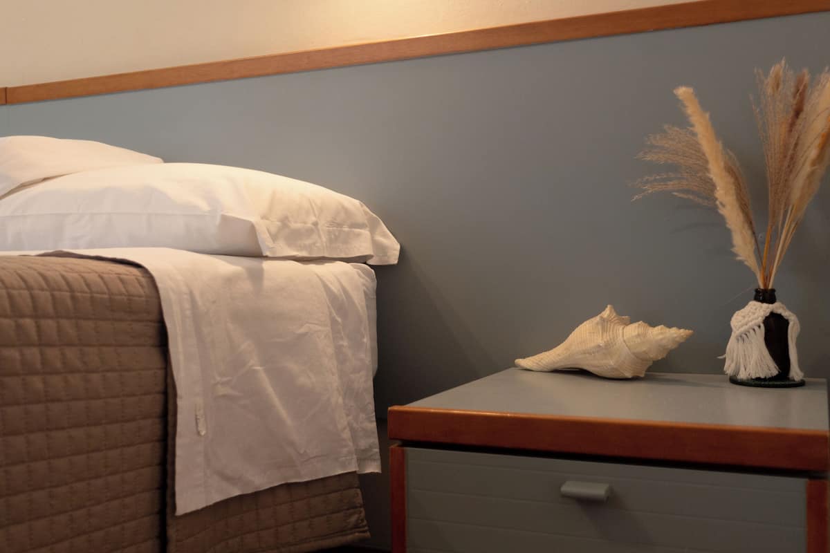 Una cama con ropa de cama blanca, una mesita de noche con una concha decorativa y hierba seca de pamapas, y un cajón con pomo.
